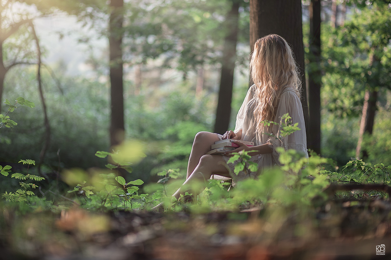 Dziewczyna siedzi w lesie na pniu, z książkami w ręku i patrzy w dal