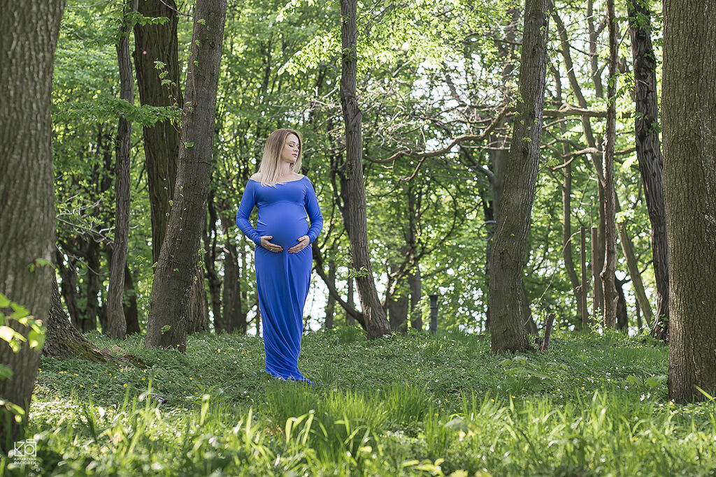 Kobieta w długiej niebieskiej sukni trzyma dłonie na swoim brzuchu, sesja ciążowa