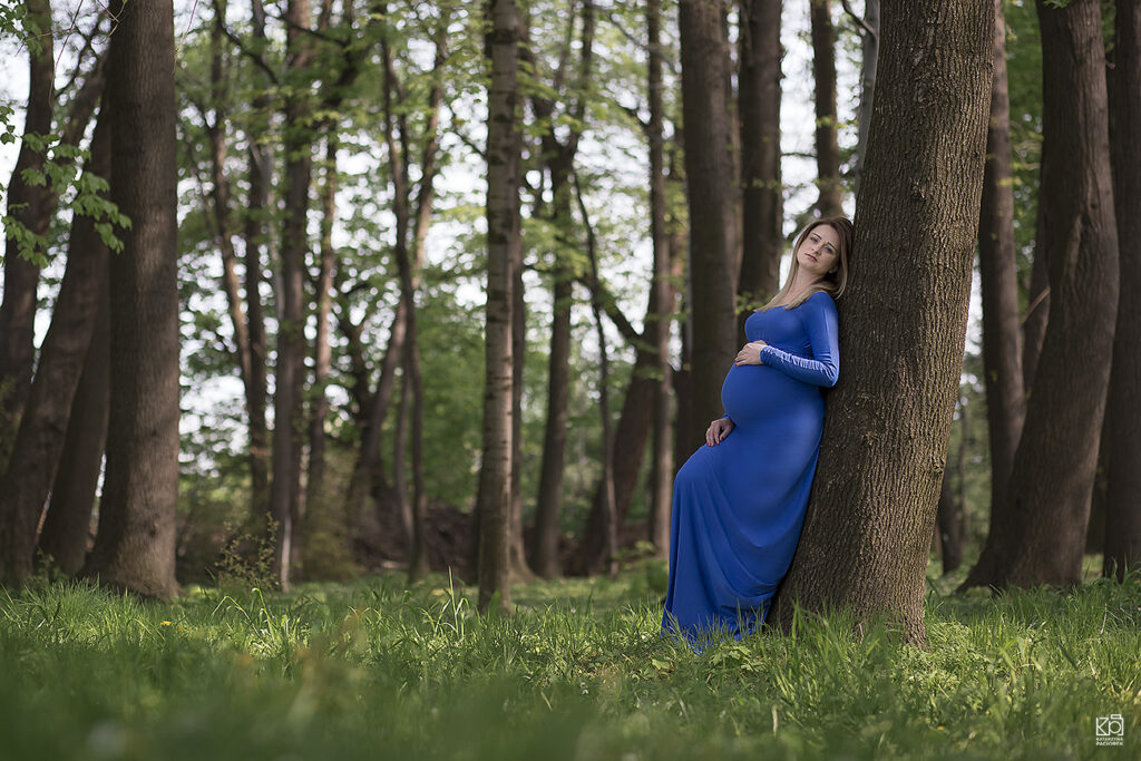 Fotografia ciążowa przedstawiająca kobietę opierającą się plecami o drzewo. Dłonie lekko położyła na swój brzuch.