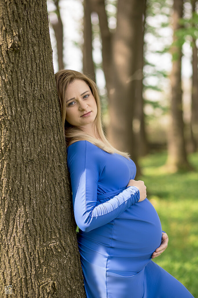 Fotografia ciążowa, kobieta oparta plecami o drzewo. Trzyma dłonie na brzuchu, wzrok zwrócony ma prosto w obiektyw