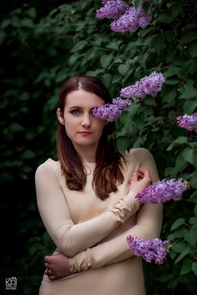 Kobieta w beżowej sukni stoi przy drzewie pełnym kwiatów bzu, lewe oko ma zasłonięte przez bez