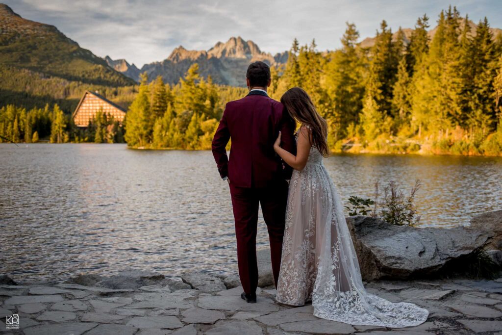 Sesja ślubna w Tatrach. Żona tuli się do meża. Oboje stoją nad brzegiem jeziora i patrzą w stronę gór