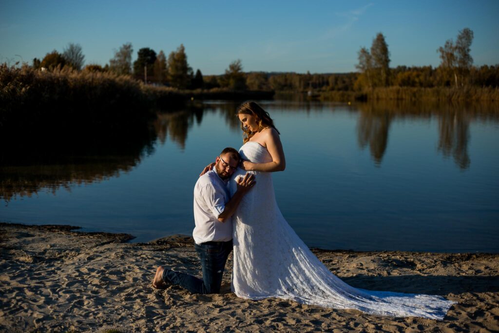 Fotografia przedstawia Karolinę i Tomka nad zalewem. Ona stoi z pochyloną głową i patrzy na Tomka, który klęczy i przytula się do ciążowego brzucha.
