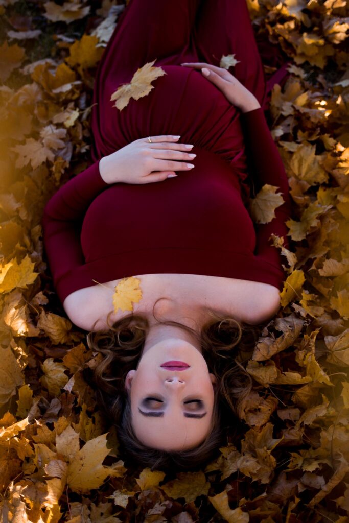 Karolina leży wśród kolorowych liści. Ma zamknięte oczy, a dłońmi obejmuje ciążowy brzuch.