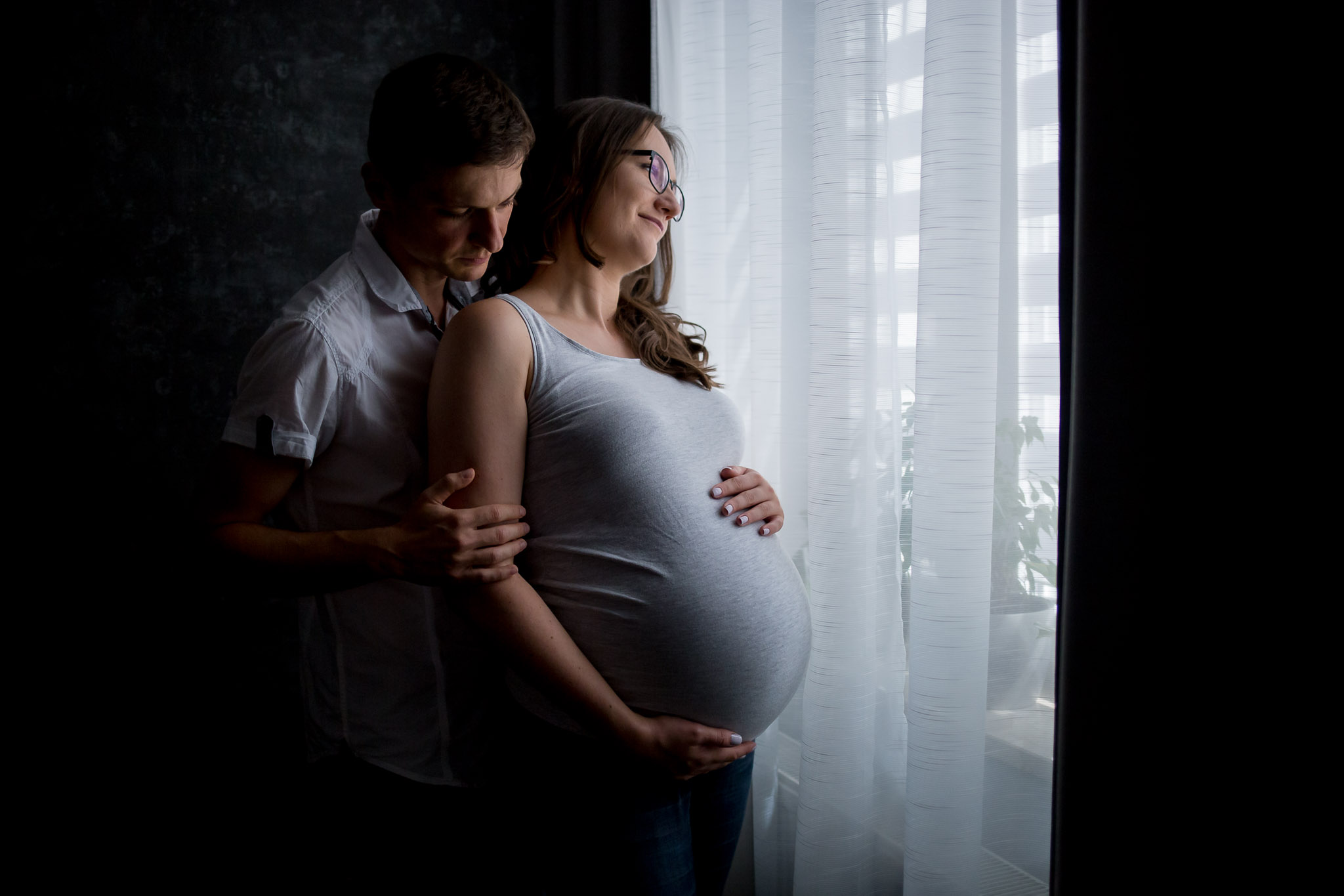 Kobieta obejmuje swój ciążowy brzuch i patrzy w okno, mężczyzna obejmuje żonę.